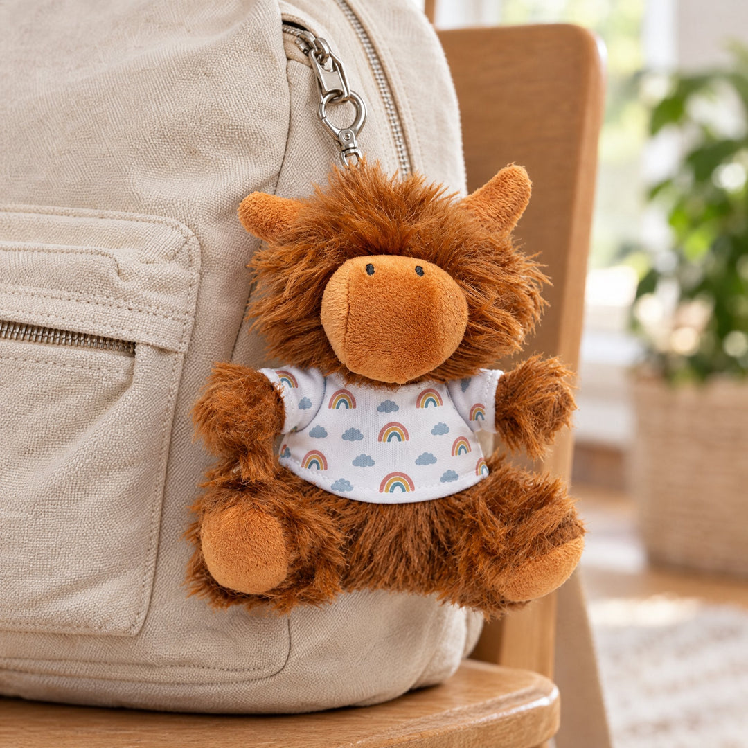 Sublimation Highland cow plush keyring printed with little rainbows and clouds, clipped to a canvas backpack